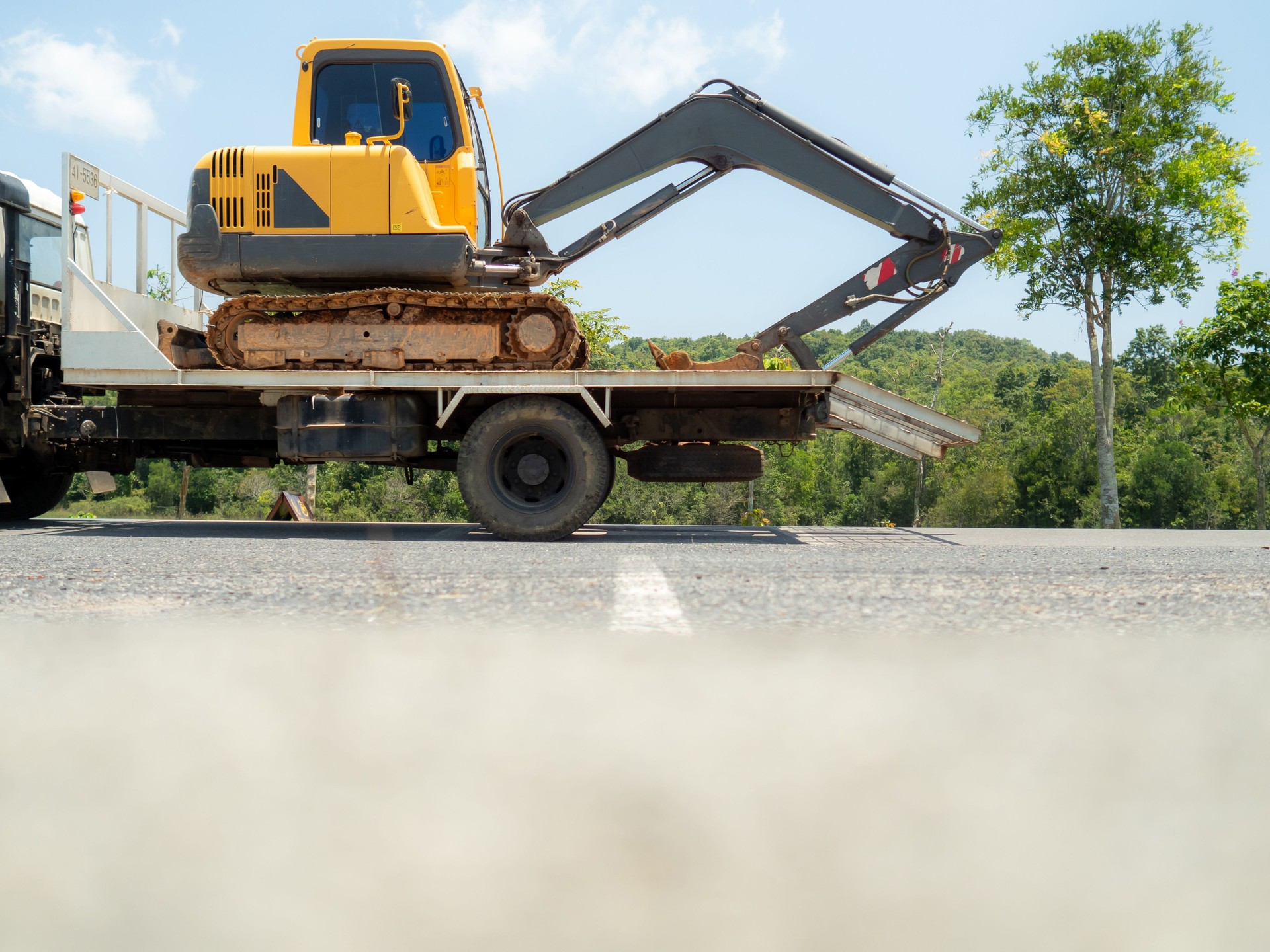 Yellow excavator being transported by flatbed truck. All trucks are running on the road. Trees along road.