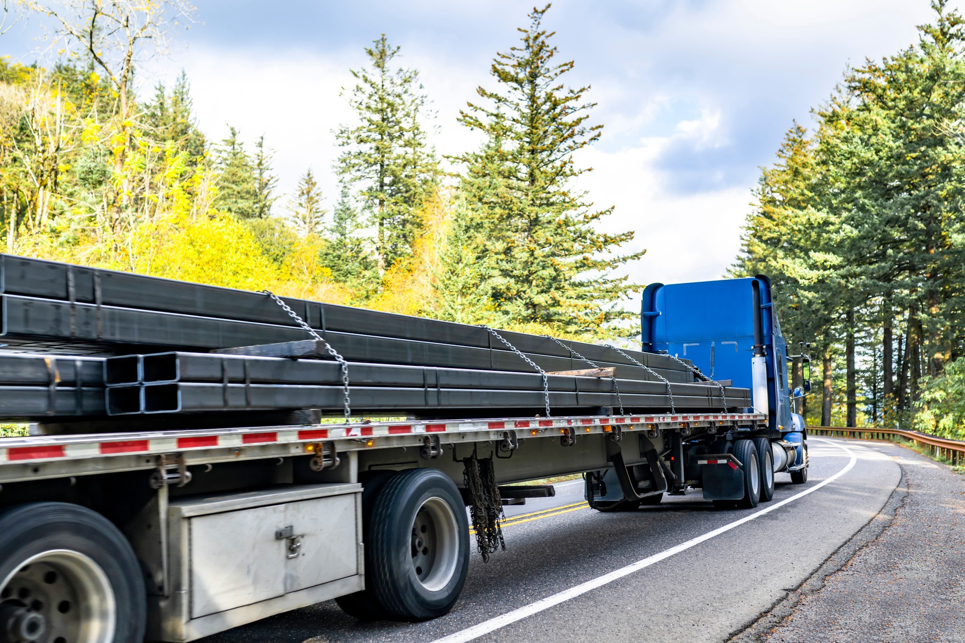 High cab blue big rig semi truck tractor transporting fastened by chains metal cargo on flat bed semi trailer running on the winding autumn highway road